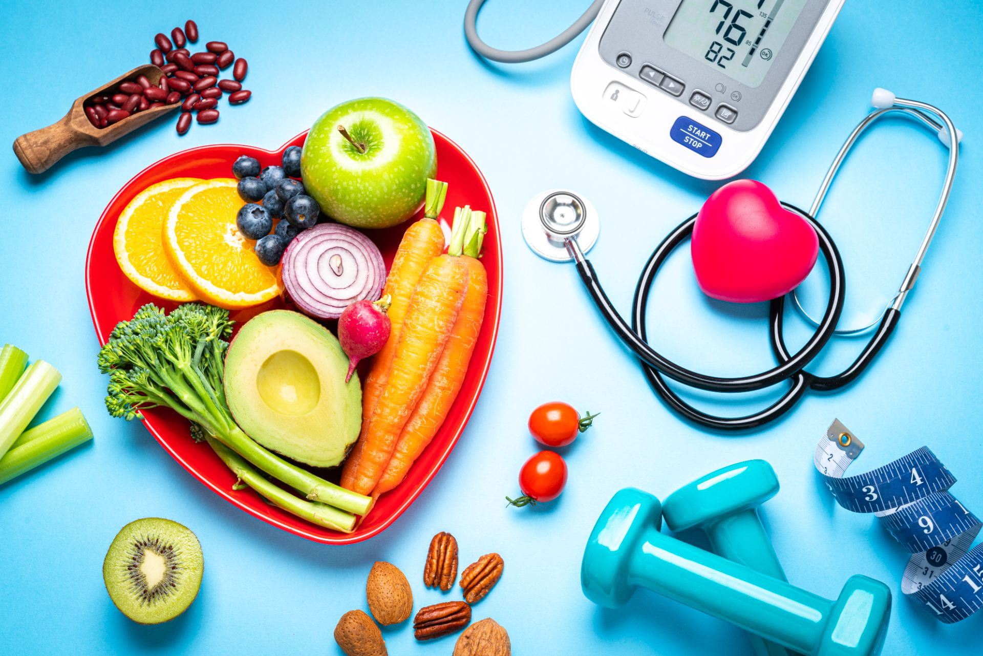 Healthy lifestyle concepts: red heart shape plate with fresh organic fruits and vegetables shot on blue background. A digital blood pressure monitor, doctor stethoscope, dumbbells and tape measure are beside the plate  This type of foods are rich in antioxidants and flavonoids that prevents heart diseases, lower cholesterol and help to keep a well balanced diet.